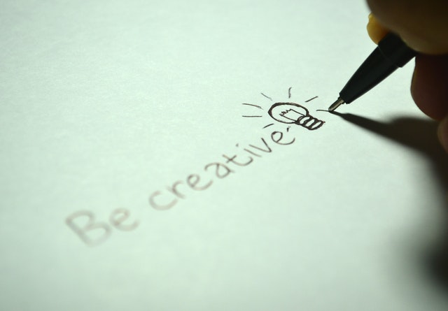 Človek píše na papier s ceruzkou nápis buďte kreatívny a kreslí žiarovku.jpg